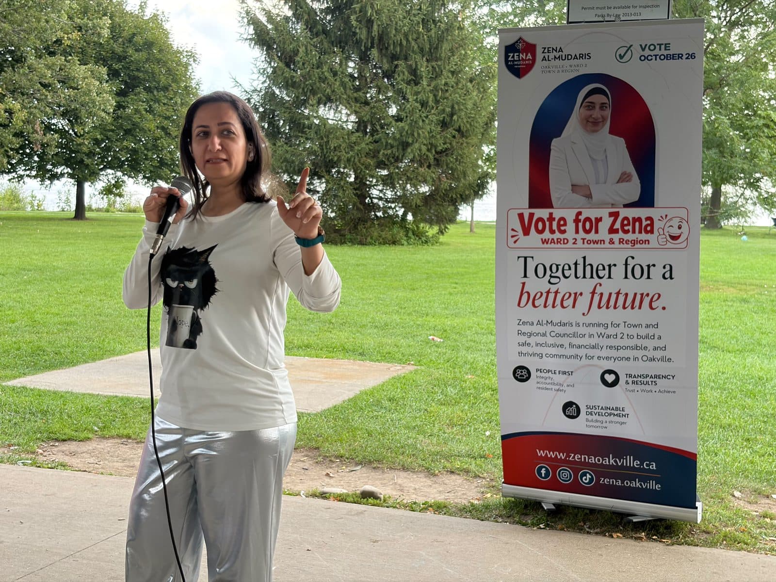 A guest in a white top speaks at the microphone beside the Vote for Zena banner