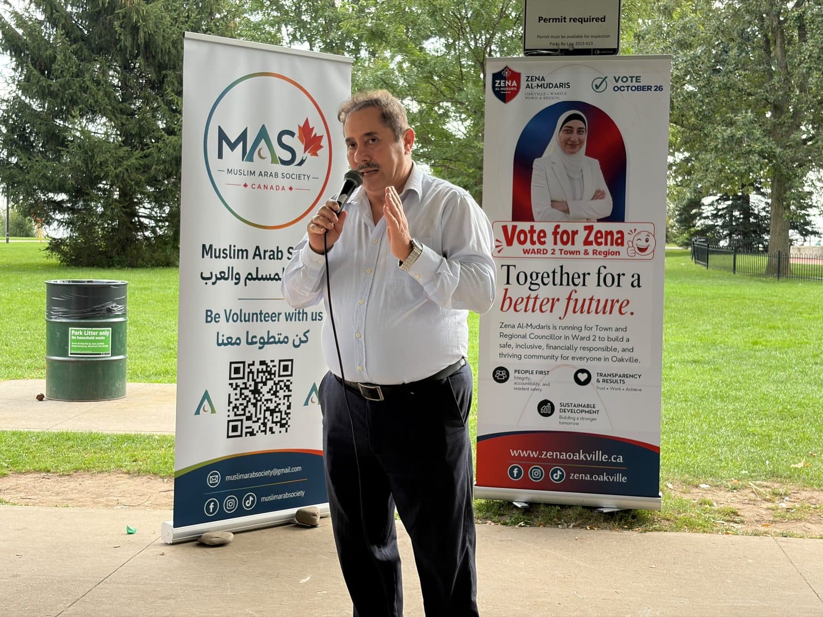 A speaker at the microphone between the Muslim Arab Society banner and the Vote for Zena banner