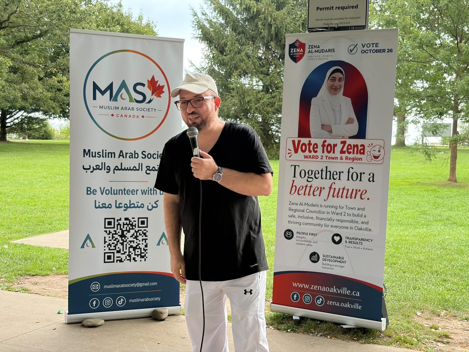 A speaker in a black T-shirt at the microphone beside the Muslim Arab Society banner