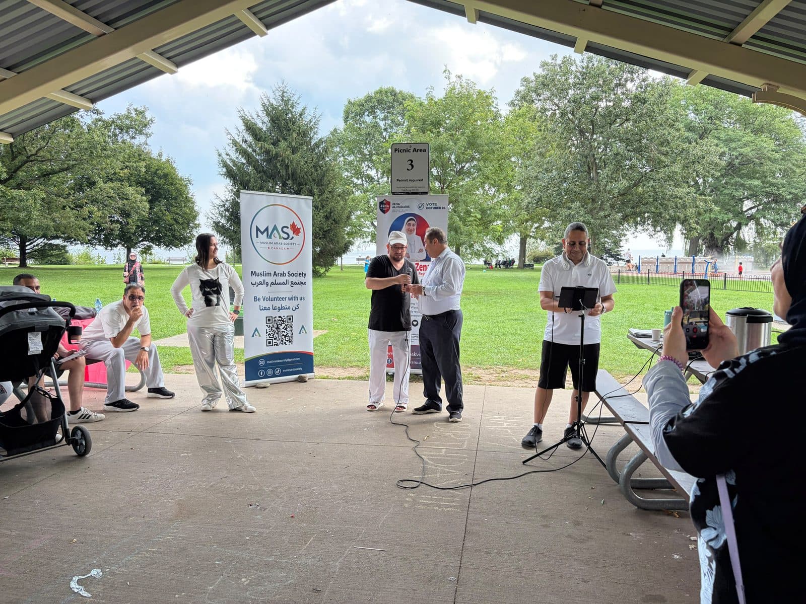 Speakers and guests at the front of the pavilion, with the lake beyond the trees