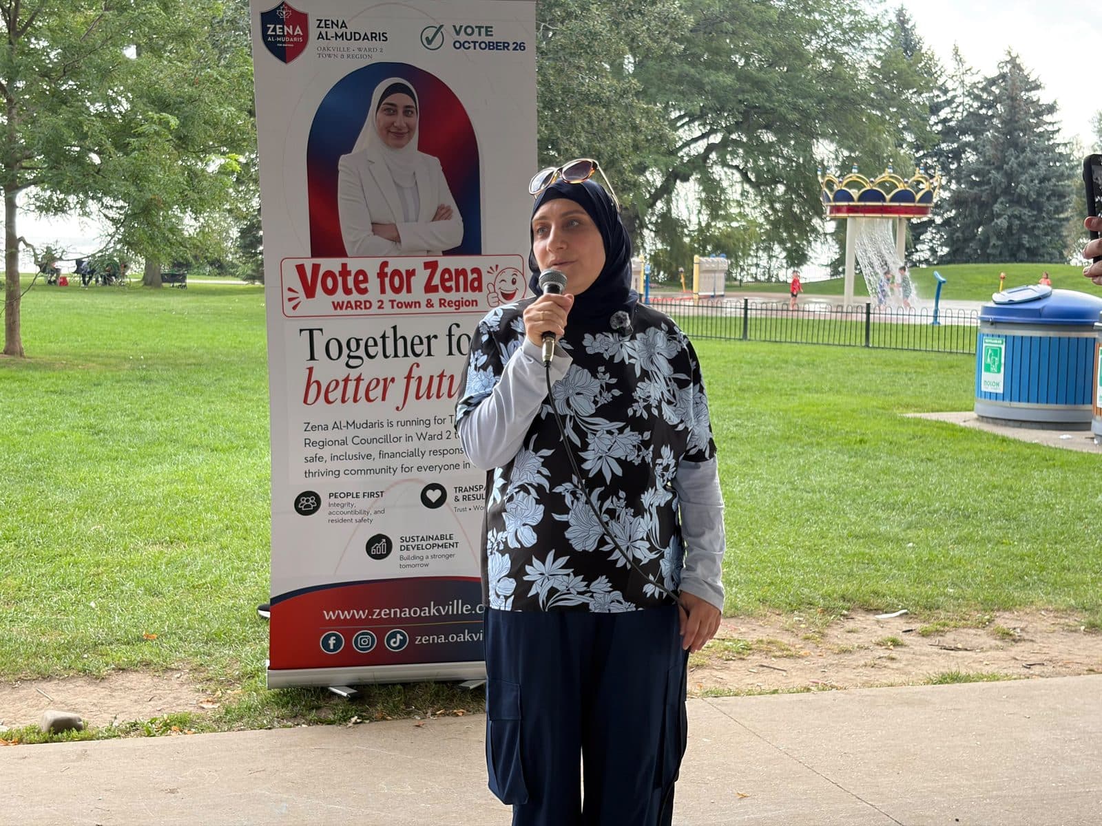 Zena speaks at the microphone beside her Vote for Zena banner under the pavilion