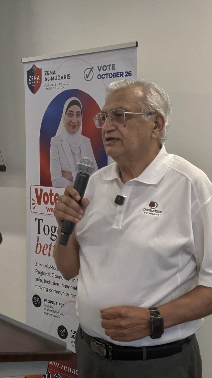 A guest in a white polo shirt speaks into a microphone beside the Vote for Zena banner