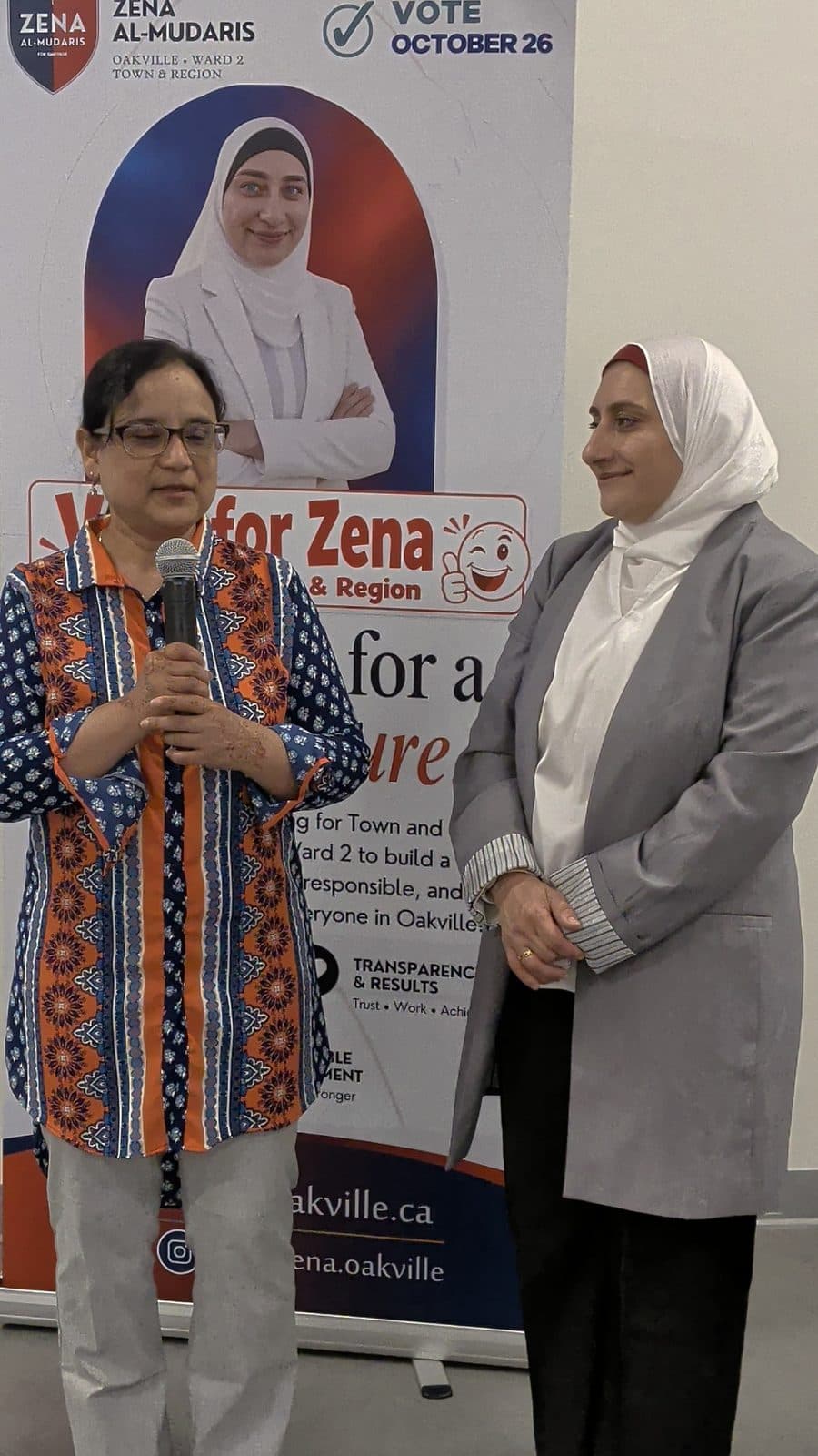A guest in a patterned tunic speaks at the microphone beside Zena