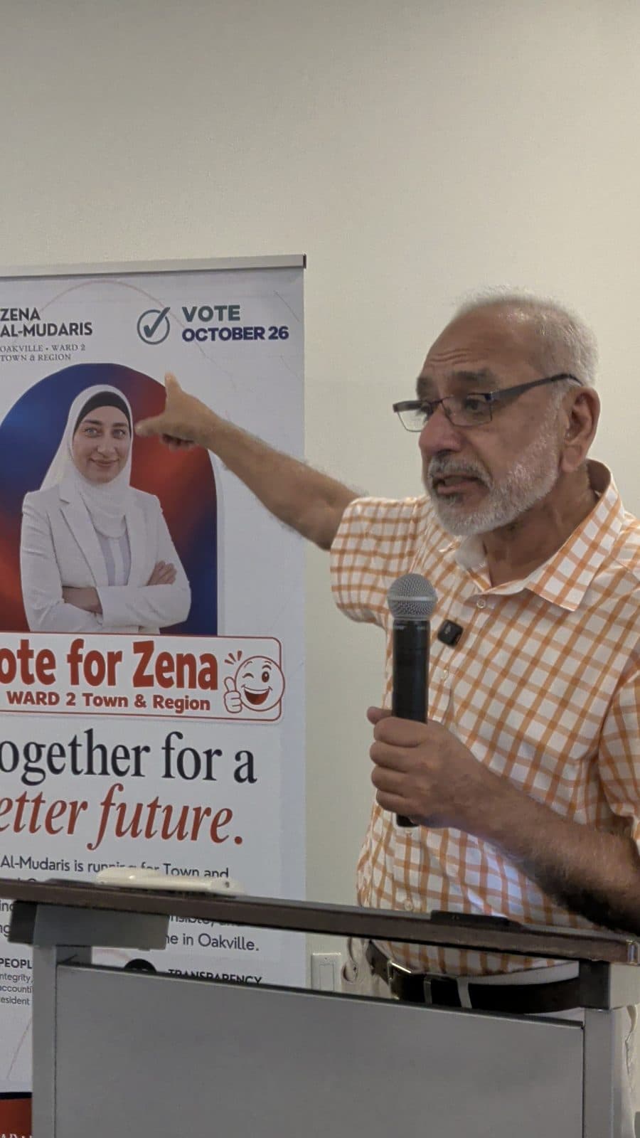 A guest at the podium points to the Vote for Zena banner as he speaks