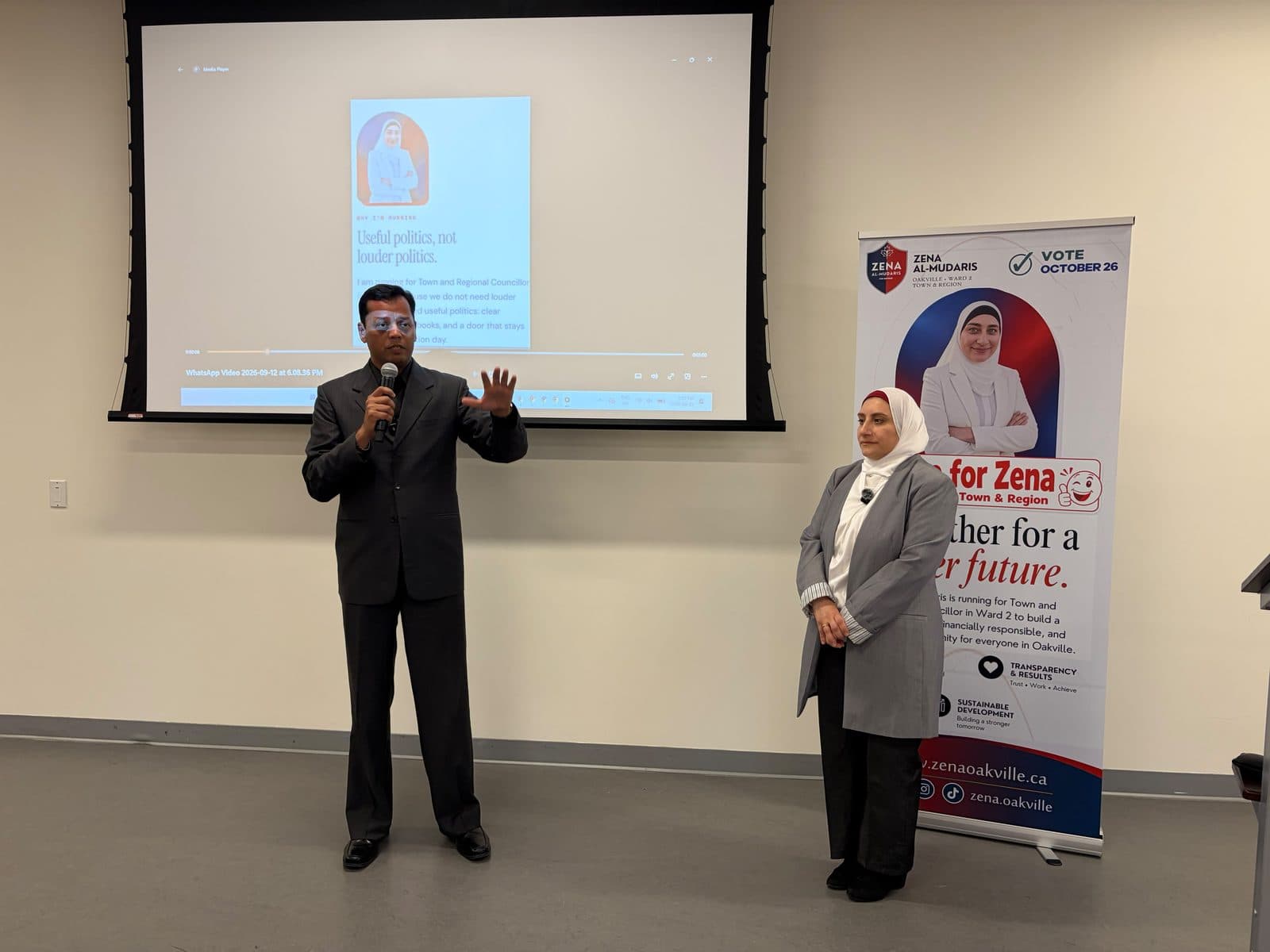 A guest speaker addresses the room while Zena stands beside her campaign banner