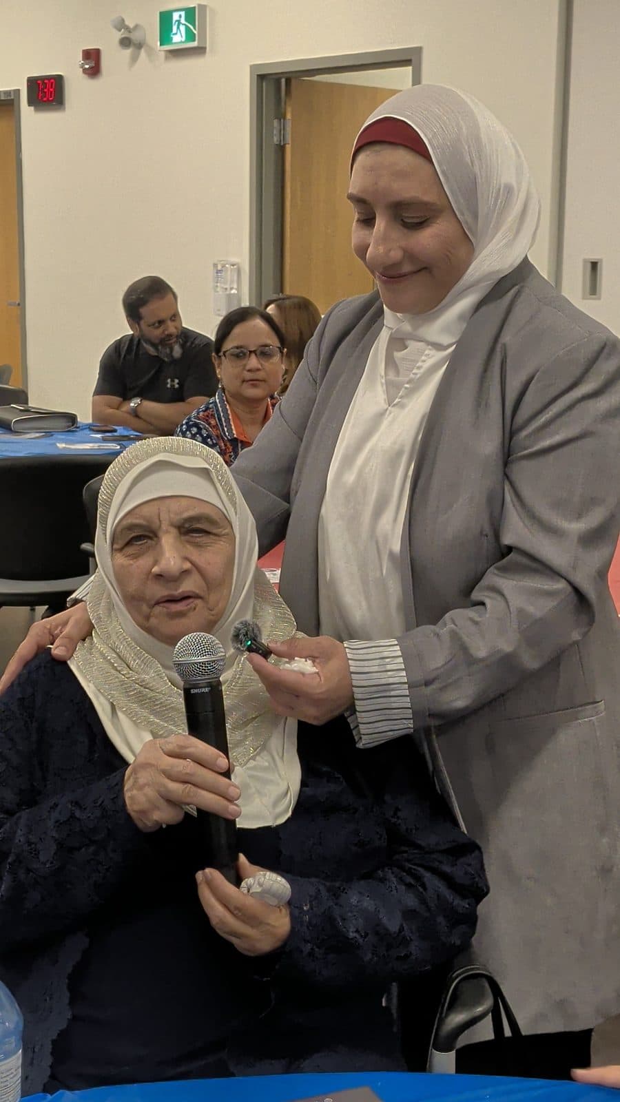 Zena stands beside a seated senior guest who holds a microphone
