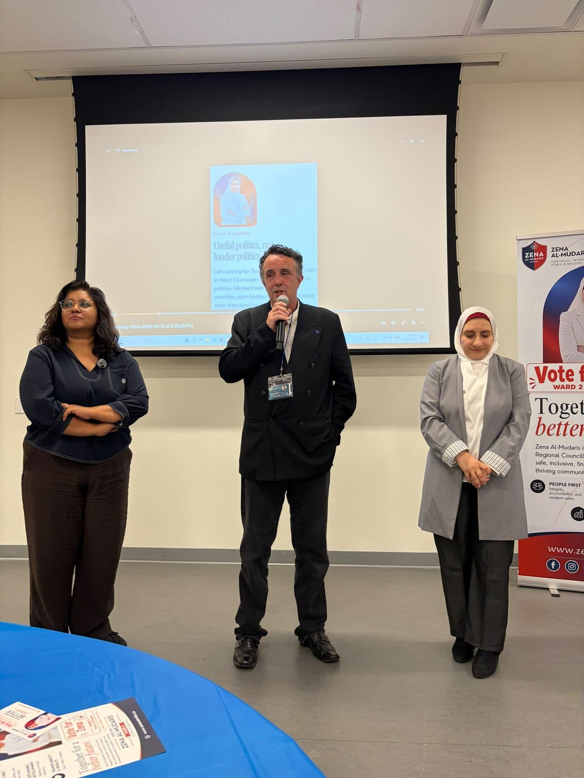 Two guests and Zena at the front of the room, with the screen showing the campaign line: useful politics, not louder politics