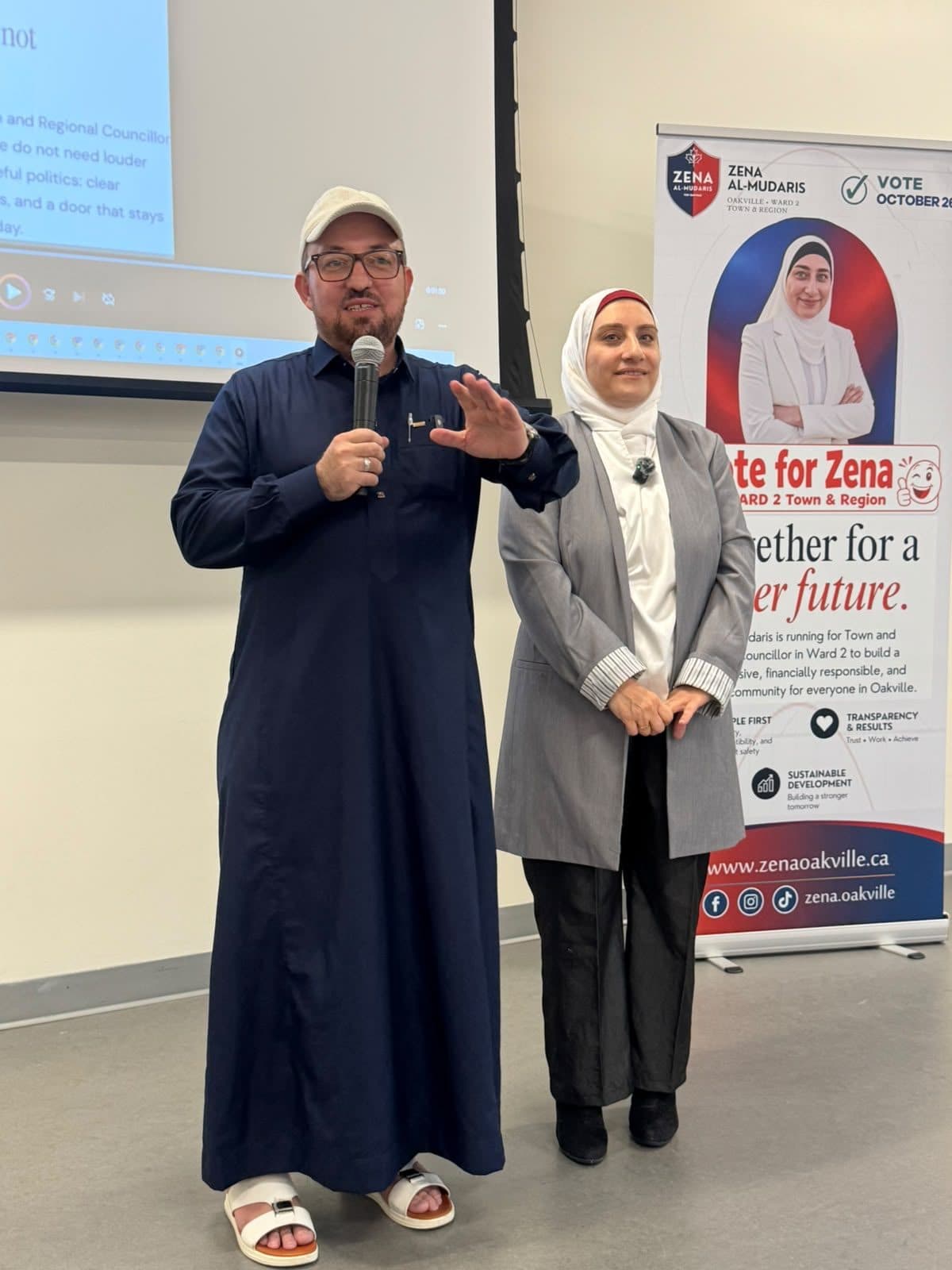 A guest in a white cap speaks at the microphone beside Zena